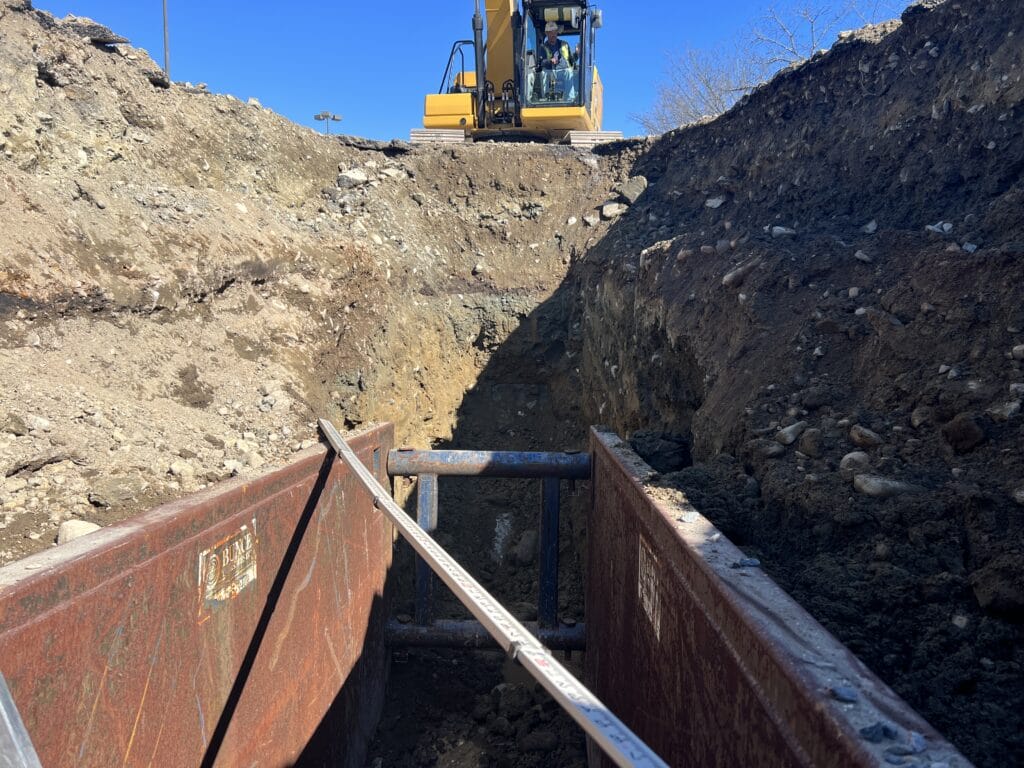 Site Work in MA for commercial development Hanover crossing in Hanover MA; trench box for utilities in trench with excavator above ground