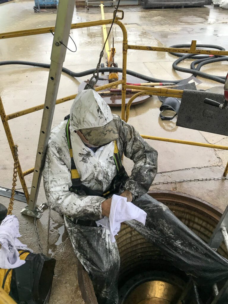 worker fully emerged for a manhole cover with safety harness, and hazmat suit, cleaning off after an inspection in a confined space.